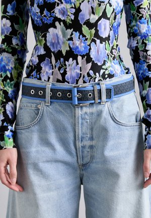 Chemise à manches longues noire à motifs floraux bleu et vert, accompagnée d'un jean bleu clair et d'une ceinture en cuir texturée bleu marine avec des accents bleu clair.