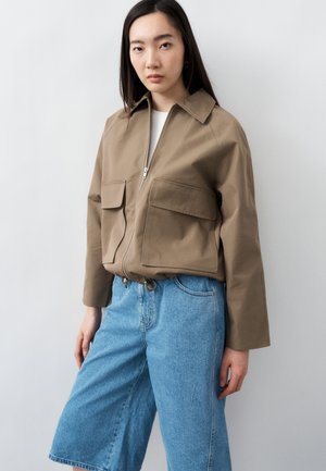 Hosszú sötét hajú nő bézs, cipzáras dzsekit visel, nagy zsebekkel és világoskék, széles szárú farmer rövidnadrágot.