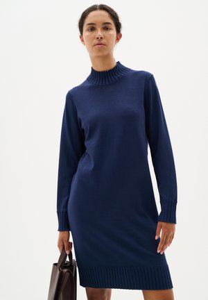 Navy knitted dress with a ribbed mock neck and ribbed cuffs, featuring a straight cut that falls above the knees. Model holds a dark brown bag.