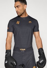 Αθλητικό τοπ με κοντά μανίκια σε μαύρο χρώμα με ανάγλυφο σχέδιο, διακοσμημένο με πορτοκαλί λογότυπο. Συνδυάζεται με μαύρα γάντια πυγμαχίας.