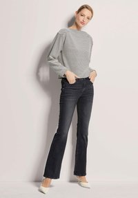 Gestreiftes Langarmhemd in Schwarz und Weiß kombiniert mit eng anliegenden, dunklen Jeans mit Schlag und weißen spitzen Absatzschuhen. Einfarbiger Hintergrund.