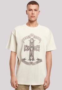 Light beige oversized T-shirt featuring a graphic design of a cross with skulls and "Guns N' Roses" text, made from cotton with a soft texture.