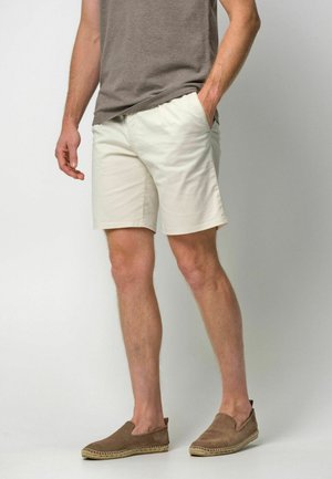 Hellbeige Shorts aus Baumwolle mit lockerer Passform, Seitentaschen und flachem Vorderteil, kombiniert mit braunen Slippern.