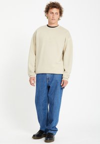 Beige fleece sweatshirt with crew neck and embroidered logo, paired with blue jeans. Black shoes complete the outfit.