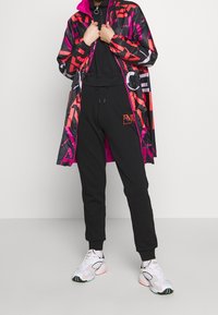 Osoba v černých teplácích a černém topu, oblečená v barevné jasně černé, červené a růžové vzorované bundě, stojící na jednobarevném pozadí.