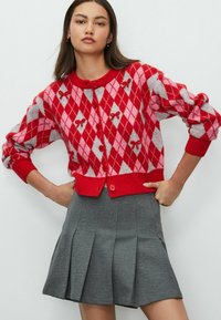 Rood en roze argyle-gepatrooneerde cardigan met grijze accenten, puff-sleeven en rode knopen; gecombineerd met een geplooide grijze rok.