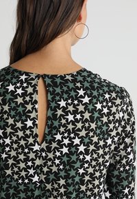 Schwarzes Kleid aus Stoff mit einem Sternenmotiv in Grün, Weiß und Grau. Verfügt über einen kleinen Schlüssellochausschnitt im Rücken und lange Ärmel.