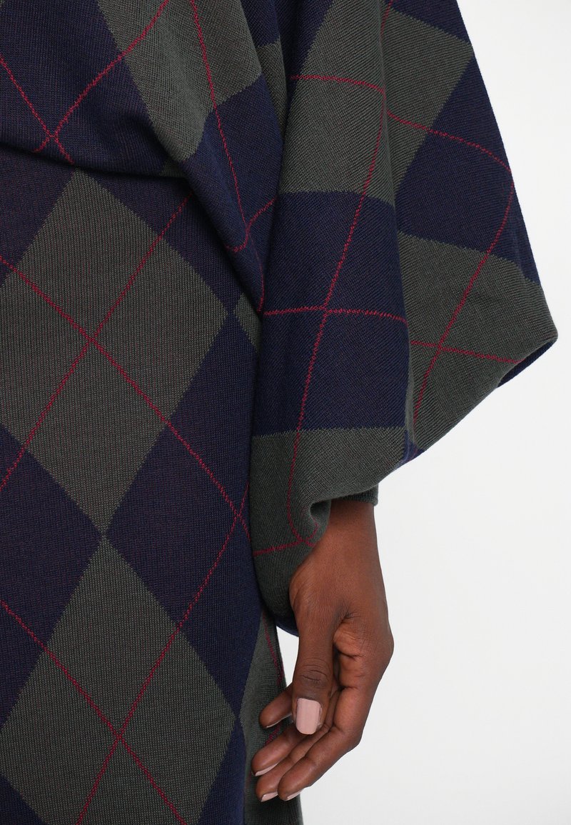 Gebreid kledingstuk met een diamantpatroon in donkerblauw, groen en rood. Zachte textuur met een gedrapeerd ontwerp, met een detail van een mouw en hand.