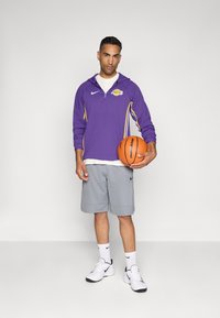 Lila Zip-Jacke mit einem Lakers-Logo, graue Shorts, weißes T-Shirt, schwarz-weiße Sneakers und einen Basketball haltend.