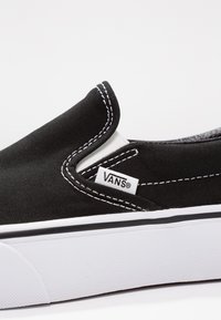 Zapatilla de lona negra de slip-on con costuras blancas y suela de goma blanca. Presenta una etiqueta lateral con el logo "VANS" y una suela texturizada.