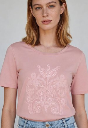 Jonge vrouw met lichtbruin haar tot op de schouders, die een roze T-shirt draagt met een ingewikkeld geborduurd bloemenpatroon en lichtblauwe spijkerbroek.