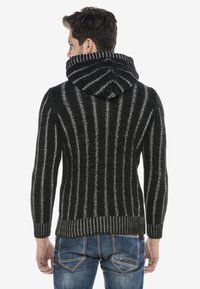 Schwarzer und grauer gestreifter Strickpullover mit Kapuze, mit gerippten Bündchen und Saum, getragen über blauen Jeans. Texturiertes Material sichtbar.