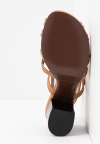 Semelle marron d’une sandale à talons hauts avec des lanières tressées couleur fauve, vue par dessous sur fond blanc.