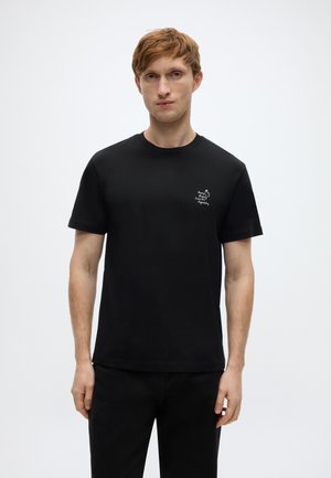 Czarny bawełniany t-shirt z krótkimi rękawami, okrągłym dekoltem i małym białym wzorem graficznym na lewej stronie klatki piersiowej. Gładka tekstura.