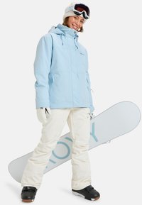Hellblaue wasserdichte Jacke mit Taschen, cremefarbene Schneehosen, schwarze Snowboardstiefel und ein weißes Snowboard mit einem blauen Logo.