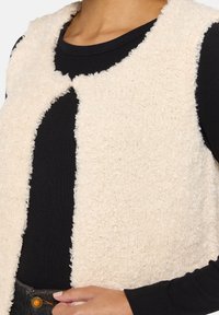 Fleece-Weste in Creme mit einer strukturierten, flauschigen Oberfläche, offenem Vorderdesign und ohne Verschlüsse, getragen über einem schwarzen Langarmoberteil.