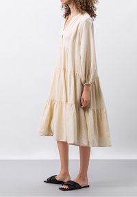 Vestido de capas en tela de color beige claro, con escote en V, mangas largas y puños elásticos, combinado con sandalias negras tipo slide.