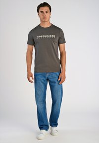 Hombre de pie vestido con una camiseta gris oscuro "Copenhagen Lindbergh", jeans azules y zapatillas blancas, frente a un fondo claro y liso.