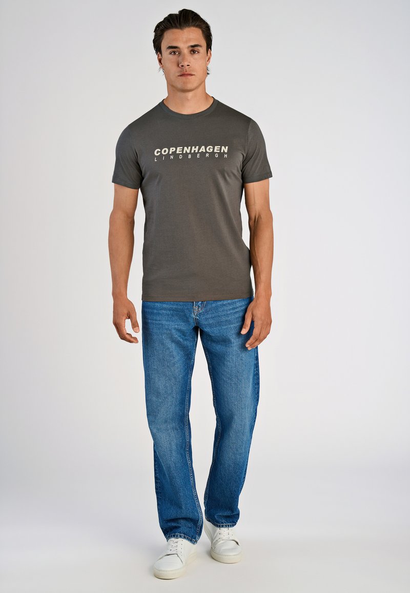 Hombre de pie vestido con una camiseta gris oscuro "Copenhagen Lindbergh", jeans azules y zapatillas blancas, frente a un fondo claro y liso.