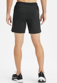 Mann in schwarzen Sportshorts und schwarzen Laufschuhen steht mit dem Rücken zur Kamera vor einem weißen Hintergrund.