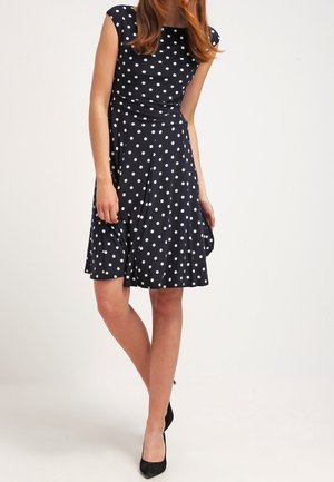Femme portant une robe noire mi-longue à pois blancs et des talons noirs, debout les jambes croisées et les mains relâchées.