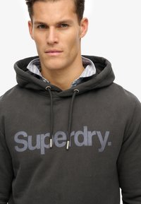 Sudadera gris oscuro con el logo "Superdry" en azul, que cuenta con una capucha ajustable y un tejido texturizado. Superpuesta sobre una camisa de rayas azul claro.