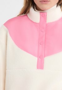 Pullover in pile color crema con collo sintetico rosa e bottoni a pressione, caratterizzato da un design a blocchi di colore con una texture morbida.