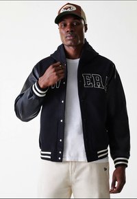 Man wearing black varsity jacket with "NEW ERA" text, white t-shirt, beige pants, and a brown and beige cap, standing against a white background.