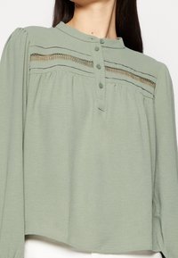 Blusa verde chiaro con scollo rotondo, patta di bottoni e dettagli in pizzo orizzontali. Maniche lunghe con spalle arricciate, texture morbida.