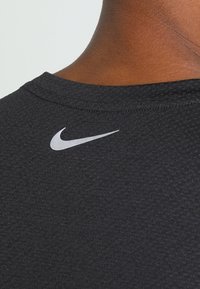 Zbliżenie na ciemną, textured sportową koszulkę z tyłu, z białym logo Nike w kształcie łuku poniżej dekoltu i widoczną skórą na szyi.