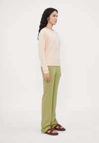Felpa pesca con scritte bianche, pantaloni verdi, sandali marroni. Il modello è in posizione laterale, evidenziando il taglio rilassato e lo stile casual dell'outfit.