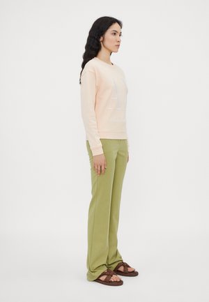 Felpa pesca con scritte bianche, pantaloni verdi, sandali marroni. Il modello è in posizione laterale, evidenziando il taglio rilassato e lo stile casual dell'outfit.