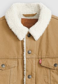 Chaqueta de denim marrón claro con un cuello de borreguito blanco, botones a presión plateados y una etiqueta roja en el bolsillo. Tejido texturizado con costuras en contraste.
