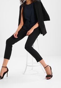 Svart skräddarsydd kavaj över en marinblå topp med dragsko i nederkant, kombinerat med svarta tajta byxor och svarta klackar med remmar och dekorativa detaljer.