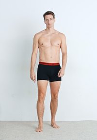 Schwarze Boxershorts mit einem roten elastischen Bund, ausgestattet mit einem figurschmeichelnden Design und weichem Material. Das Modell steht barfuß vor einem schlichten Hintergrund.
