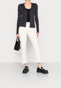 Cardigan a coste scuro con bottoni, canotta nera, pantaloni bianchi a vita alta, e mocassini neri con platforma. Accessorio: piccola borsa nera.