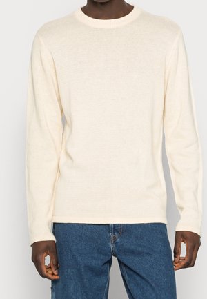 Pull long à manches longues couleur crème avec un col rond côtelé. Fabriqué en tissu doux et texturé. Associé à un jean en denim bleu.