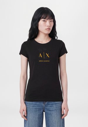 Une femme pose avec un T-shirt noir Armani Exchange orné d’un logo doré et un jean bleu sur un fond uni clair.