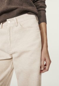 Light-colored corduroy pants with vertical stripes, front pockets, and a button closure, paired with a brown knit sweater.