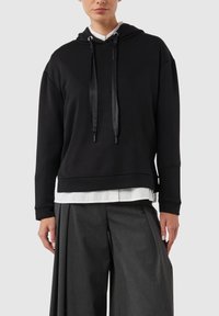 Czarna bluza z kapturem z satynowymi troczkami, wykonana z gładkiej tkaniny. Charakteryzuje się krótkim krojem i jest noszona na białej koszuli.