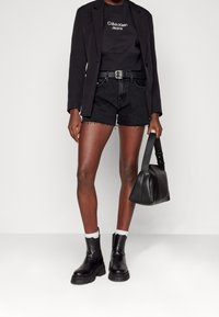 Person med svart blazer, svart Calvin Klein sweatshirt, svarta denimshorts, kraftiga svarta stövlar med vita strumpor, som håller en svart handväska.