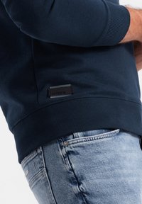 Mörkblå sweatshirt med ribbade manschetter och nederkant, med en svart etikett på sidan. Bärs med ljusblå slitna jeans.