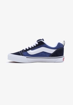 Zapatilla baja en azul marino y negro con raya lateral blanca, cordones blancos, suela blanca y tirador en el talón.