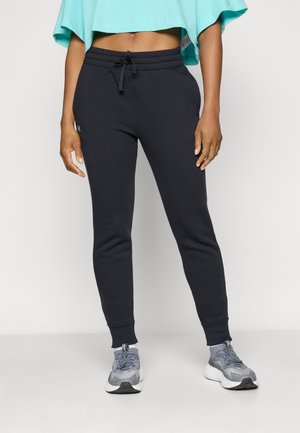 Femme portant un pantalon de jogging noir à cordon avec des poignets côtelés et des chaussures de sport grises, debout devant un fond clair uni.