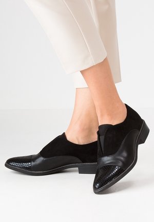 Czarne damskie buty na niskim obcasie, wykonane ze skóry i zamszu, z fakturowanymi noskami, noszone z jasnobeżowymi, krótkimi spodniami na białym tle.