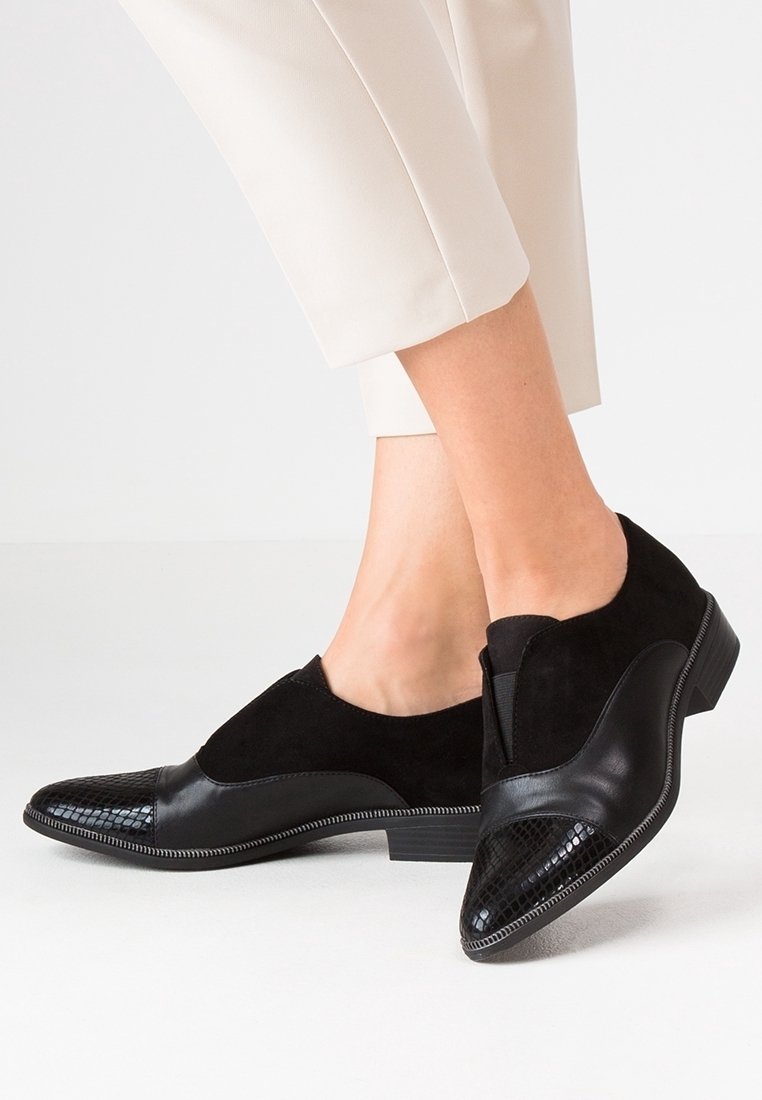 Chaussures pour femmes en cuir et suède noirs à petits talons, avec bouts texturés, portées avec un pantalon court beige clair sur fond blanc.