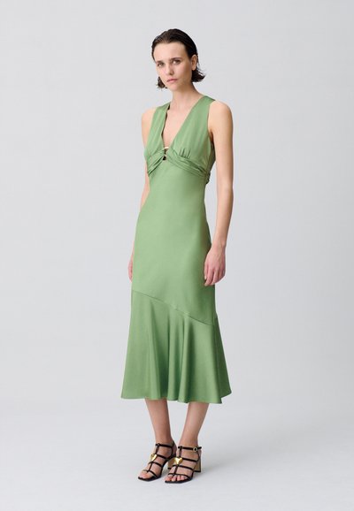 Robe en satin vert avec un décolleté en V profond, des détails froncés au niveau de la poitrine et un ourlet évasé. Sandales noires à lanières avec des accents dorés.
