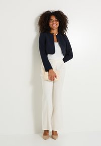 Blazer court bleu marine à col rond, associé à un haut blanc et un pantalon large crème. Tenant une pochette texturée beige clair.