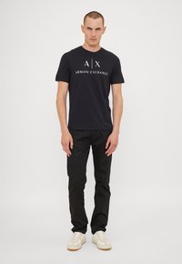 Juodas marškinėliai su balta "Armani Exchange" logotipu, trumpomis rankovėmis, derinami su juodomis kelnėmis ir baltomis sportbačiais, stovintis prieš šviesią foną.