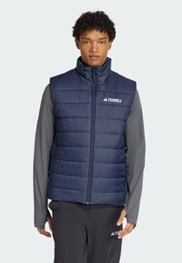 Marineblå puffer vest med vertikal quiltet tekstur, høj krave og lynlås lukning. Har et hvidt "TERREX" logo på venstre bryst.
